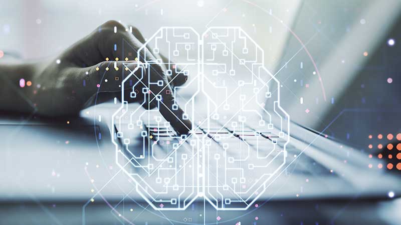 Uma mão a tocar no teclado de um computador portátil, com a imagem gráfica de um cérebro em marca de água sobre a fotografia